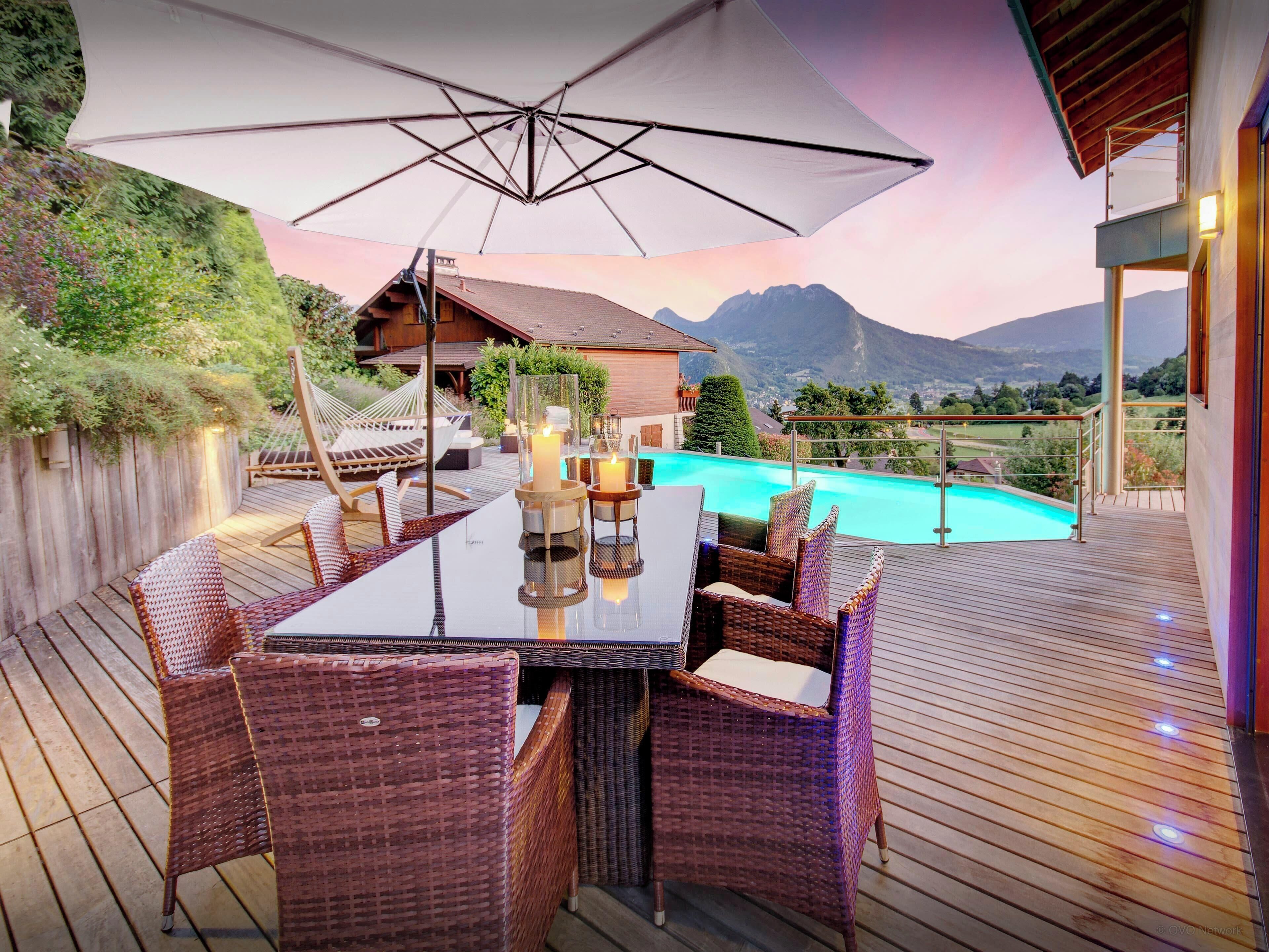 A wooden deck with a dining table and wicker chairs, overlooking a swimming pool and mountains.