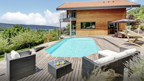 Ein modernes Haus mit Holzterrasse, Swimmingpool, Gartenmöbeln und Blick auf die Berge.