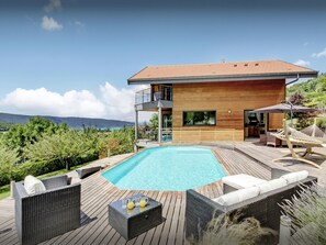 Una casa moderna con terraza de madera, piscina, mobiliario exterior y vistas a las montañas.