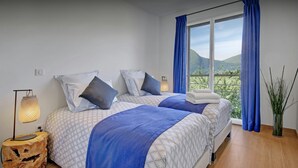 Ein Schlafzimmer mit zwei Betten, einem Nachttisch, einer Lampe und Blick auf Grün durch das Fenster.