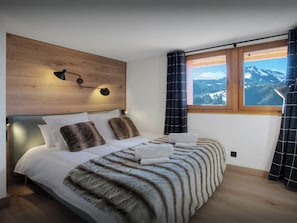 Un dormitorio con cabecera de madera, un ventanal con vistas a la montaña y una cama con sábanas blancas y una manta de piel.