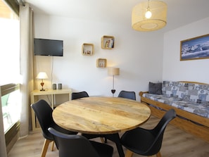 A cozy room with a round wooden table, four chairs, a sofa with a patterned cover, a television mounted on the wall, and a window with curtains. - Renovated 5 pers. apartment with south balcony, station center (La Toussuire)
