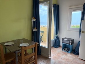A small room with a table set for two, a door leading to a balcony, a small TV stand, and a window with a view.