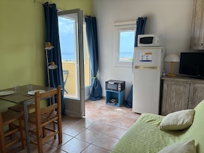 A compact room with a dining table, chairs, a small kitchenette with a microwave and refrigerator, a television, and a view through a door.
