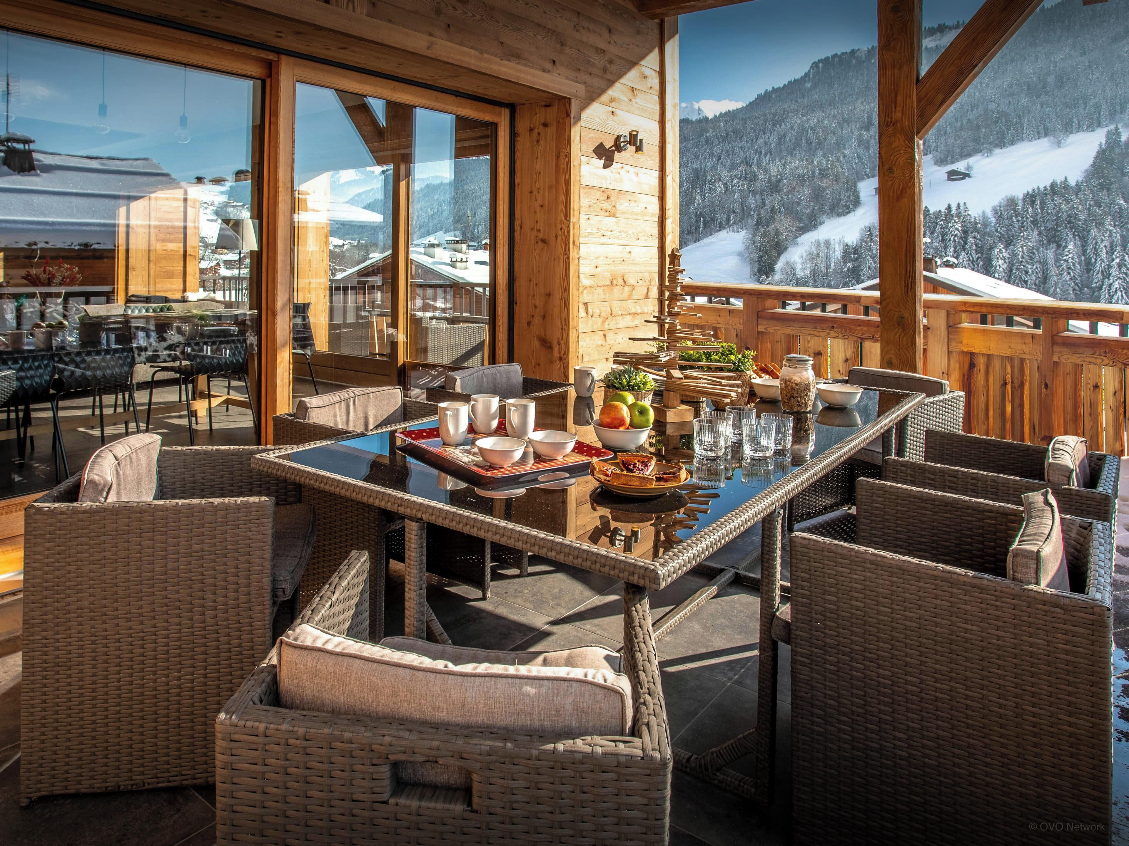 Een houten terras met rieten meubels en een glazen tafel, met uitzicht op een besneeuwd berglandschap.