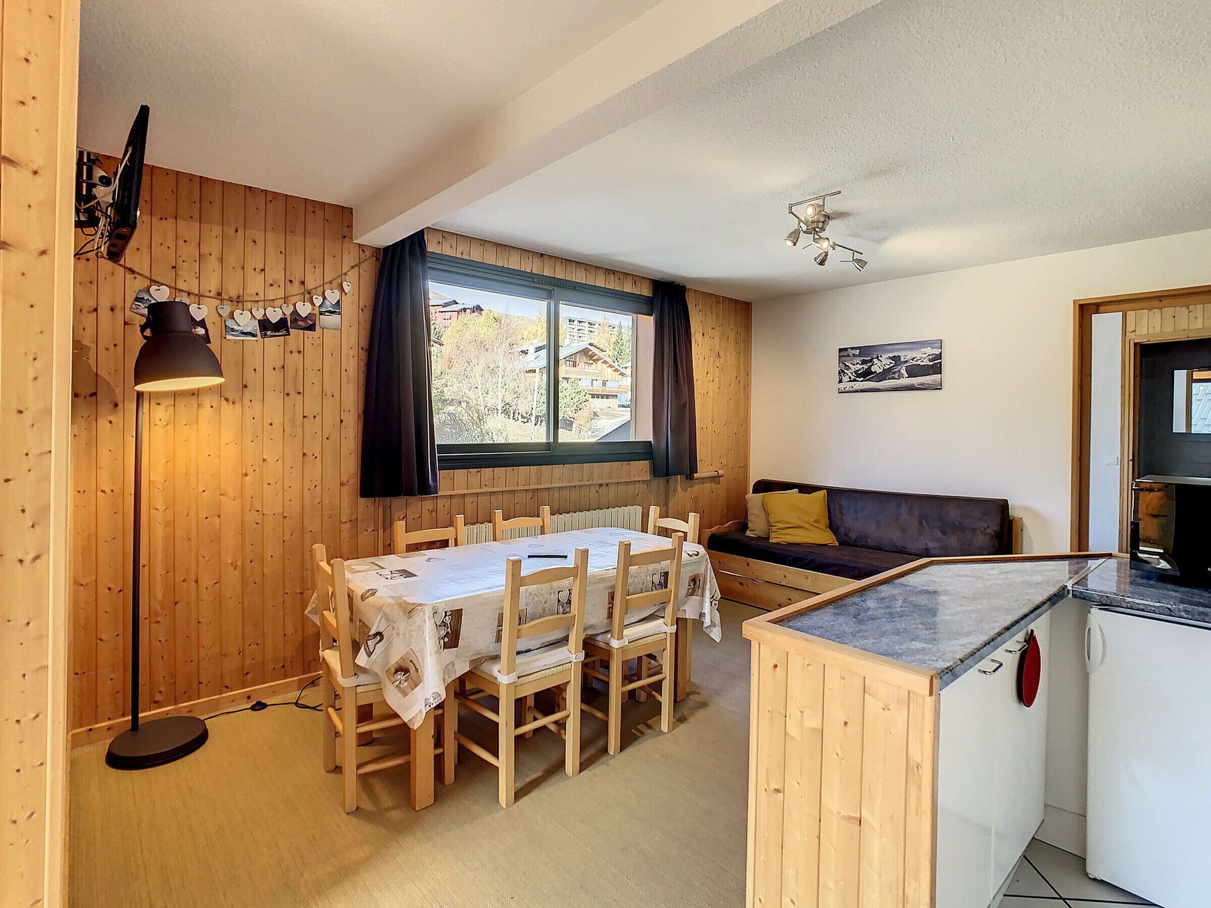 A cozy room with a wooden dining table, chairs, a sofa, a small kitchen area, and a window with curtains.
