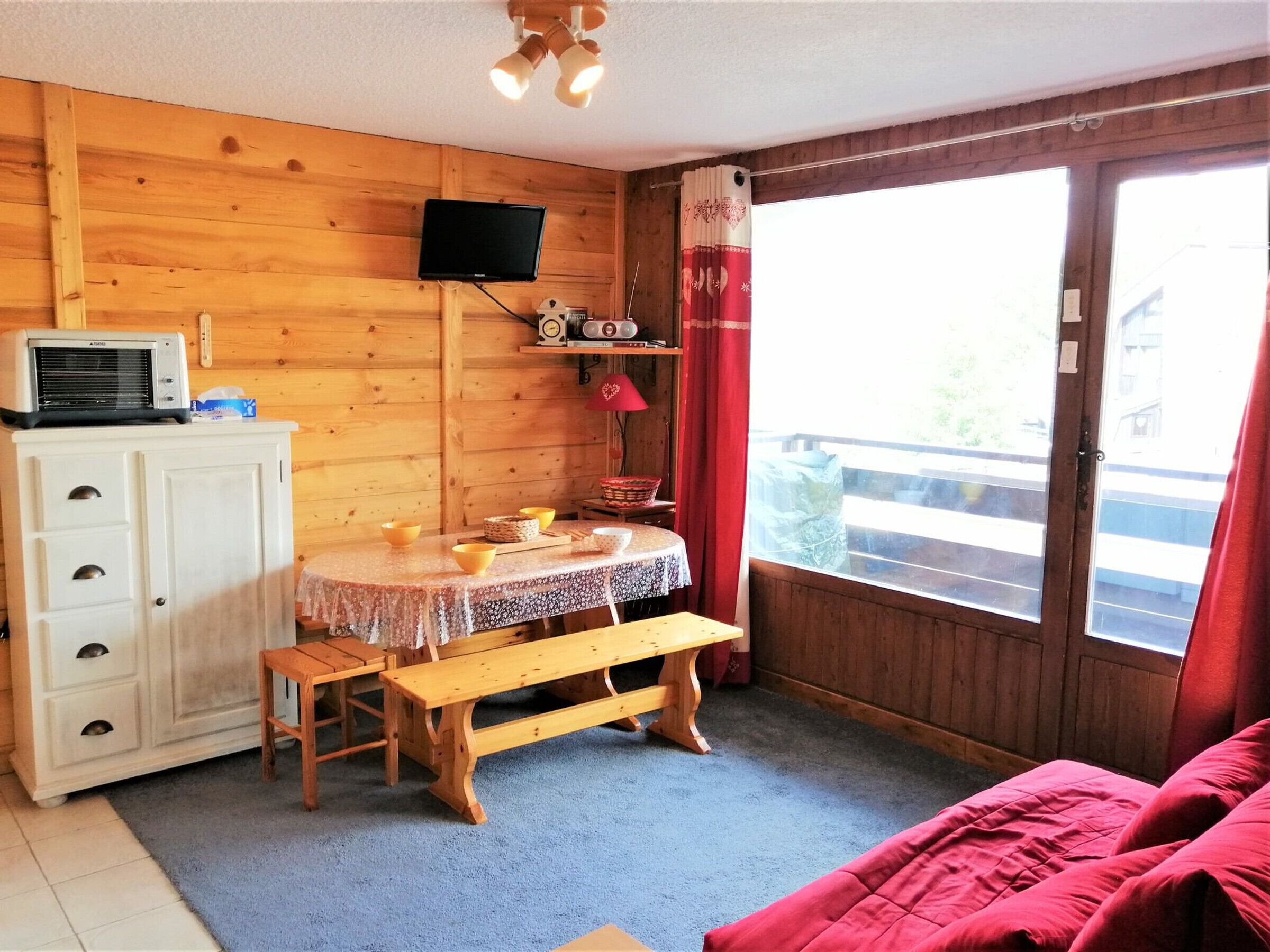 A cozy room with a wooden interior, a round table with a lace tablecloth, a bench, a white cabinet with drawers, a television mounted on the wall, and a large window with curtains.