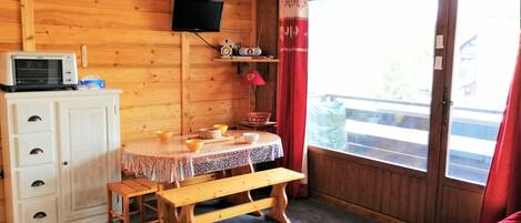 A cozy room with a wooden interior, a round table with a lace tablecloth, a bench, a white cabinet with drawers, a television mounted on the wall, and a large window with curtains.