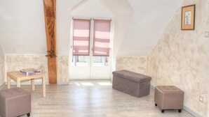 Une pièce mansardée spacieuse, avec un plafond en pente, une poutre en bois, une petite table avec un coffre, deux bancs rembourrés et une grande fenêtre avec des stores.