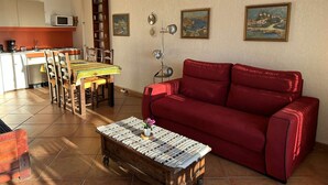 Un espace de vie compact comprenant un canapé rouge, une table basse en bois, une table à manger avec des chaises, un coin cuisine avec un four à micro-ondes et deux tableaux encadrés accrochés au mur.
