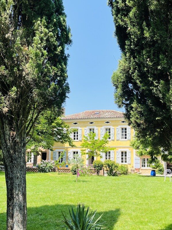 Un bâtiment à deux étages, doté d’une façade jaune et de volets blancs, entouré d’une pelouse bien entretenue et d’arbres.
