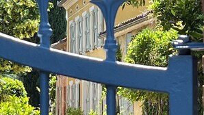 Ein blaues Metalltor mit verziertem Knauf, ein gelbes Gebäude mit weißen Fensterläden und üppiges Grün im Hintergrund.