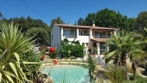 Una casa de dos pisos con piscina, rodeada de plantas y árboles tropicales.