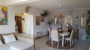 A living room with a sofa, a dining table with chairs, a hanging light fixture, and a kitchen area with a stove.
