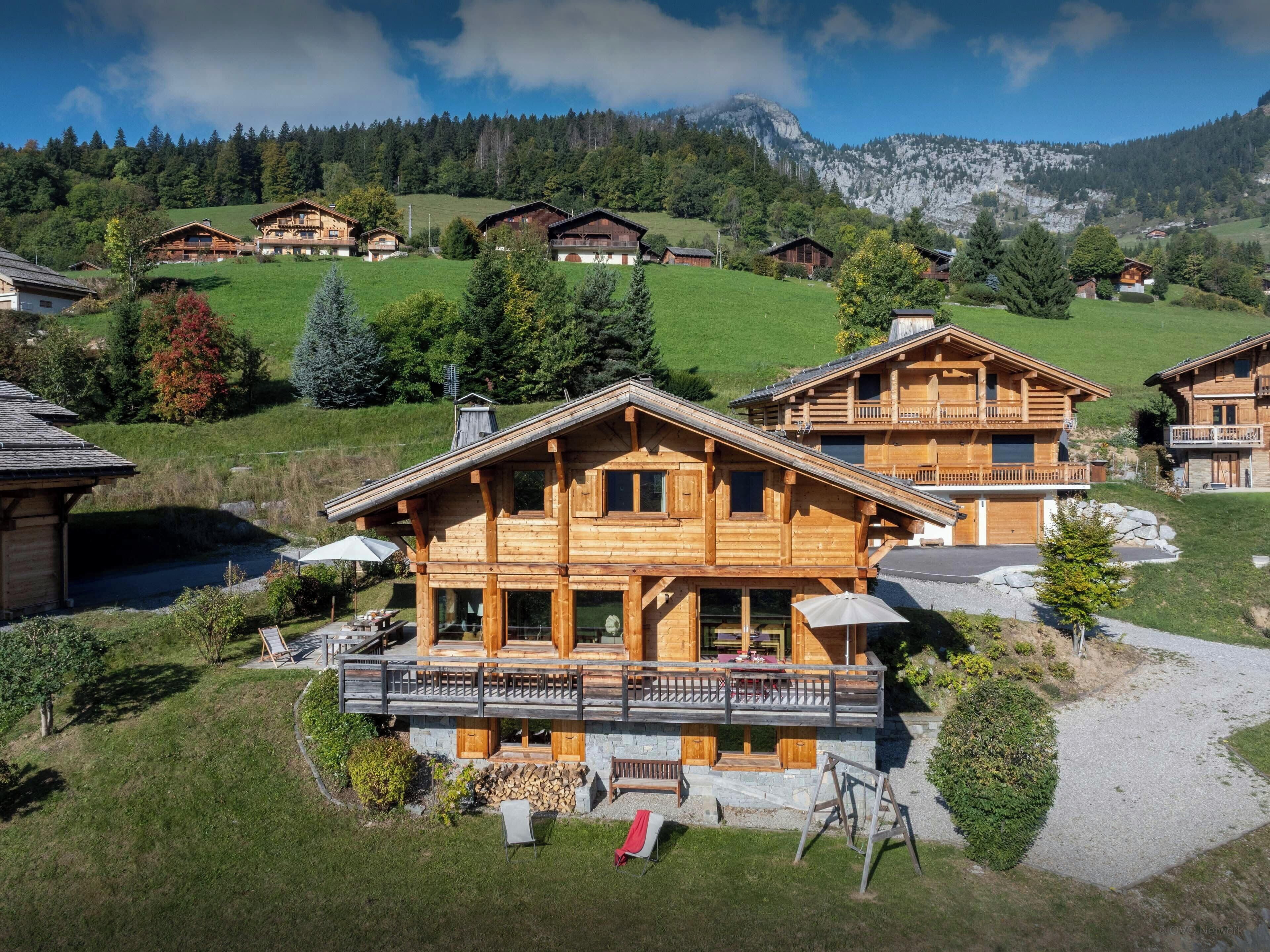 A wooden chalet with a balcony, surrounded by a lush green landscape and mountains in the background.