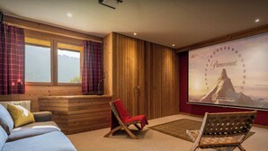 A cozy room with a wooden cabin-style interior, a large window with curtains, a red folding chair, a sofa, and a wall-mounted projector screen displaying a Paramount advertisement.