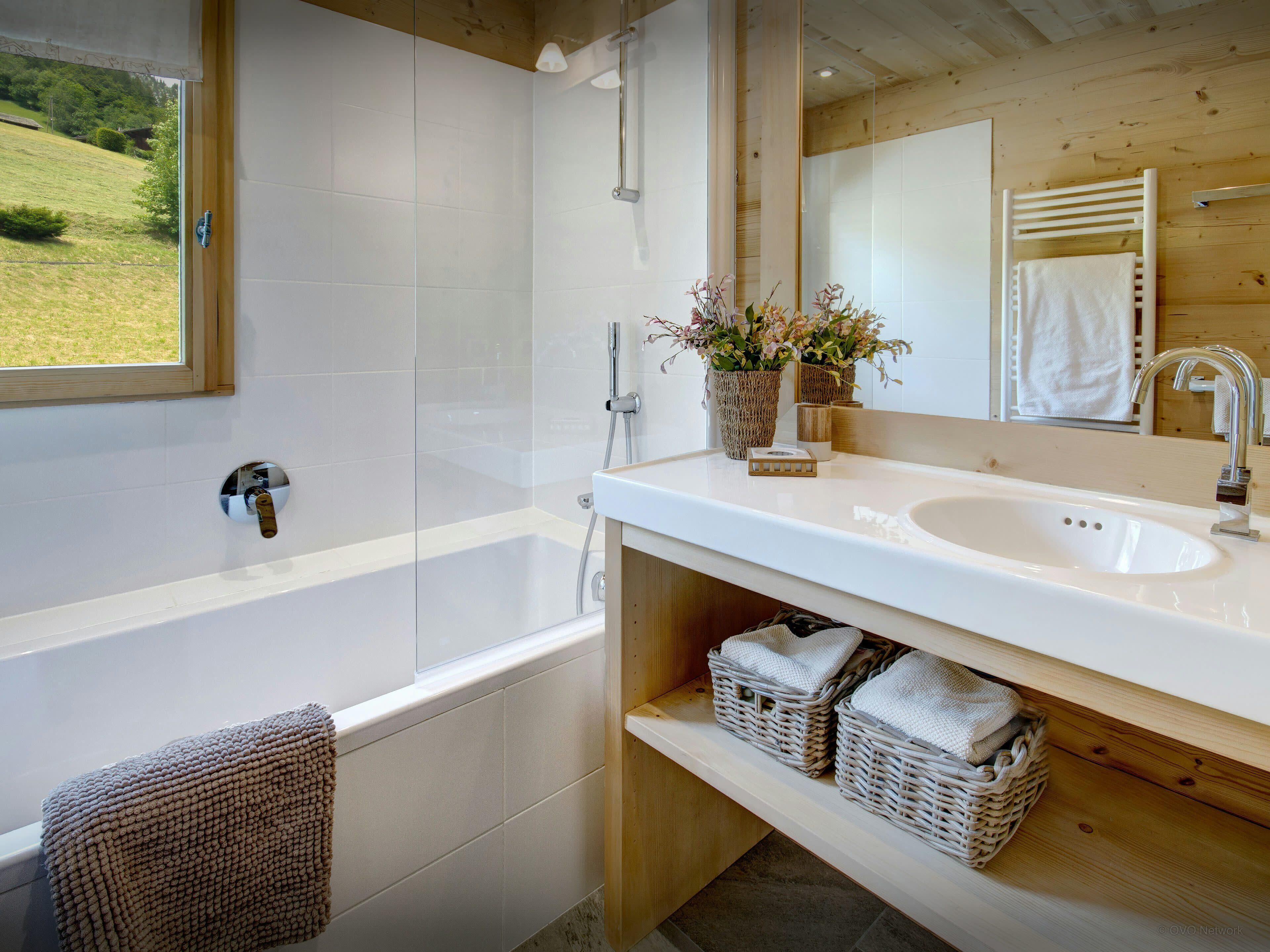 Une salle de bain moderne avec une baignoire, un lavabo et un grand miroir. On trouve des paniers en osier sous le lavabo et une serviette accrochée au mur.