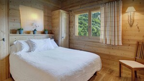 A wooden cabin bedroom with a large bed, a chair, a window with curtains, and a shelf with plants.