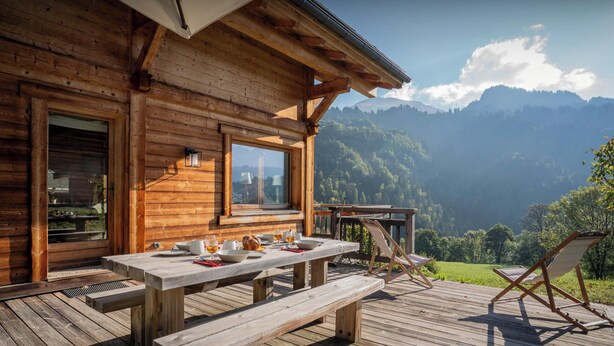 A wooden cabin with a wooden deck, a table set for tea, and a view of mountains.