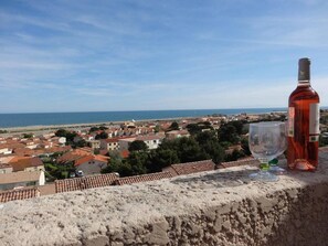 Uma vila costeira com edifícios de telhados em tons de terracota, céu azul límpido e uma garrafa de vinho tinto numa saliência.