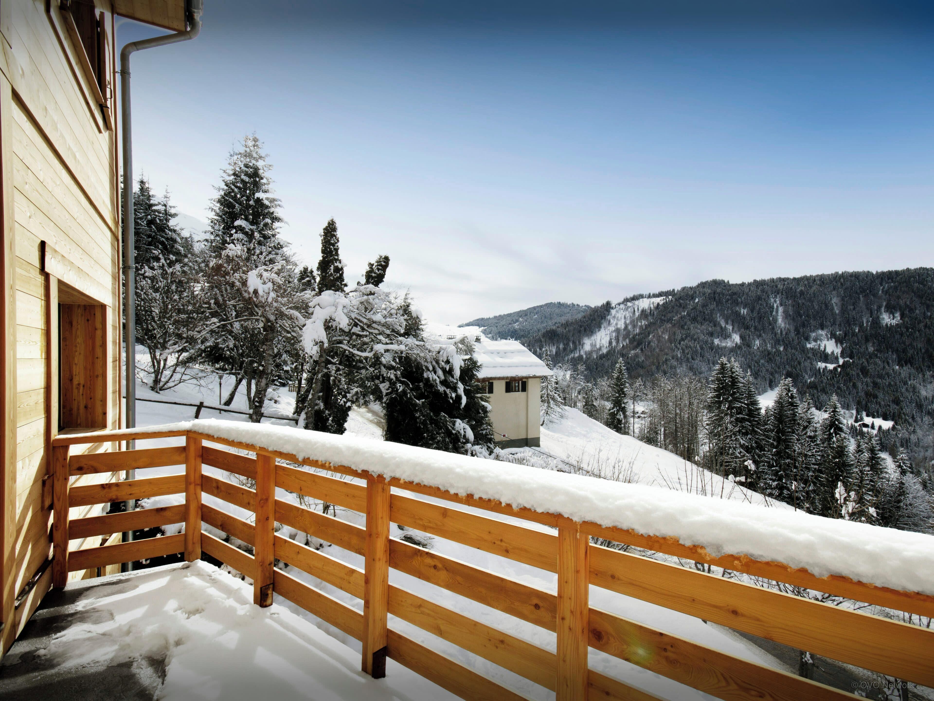 Un balcon en bois aux balustrades recouvertes de neige, offrant une vue sur un paysage montagneux enneigé.