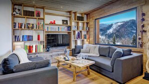 A cozy living room with a large bookshelf, a fireplace, a sofa, and a large window with a mountain view. - Chalet Charmar - secluded ski-in ski-out chalet for 12 - OVO Network (La Clusaz)