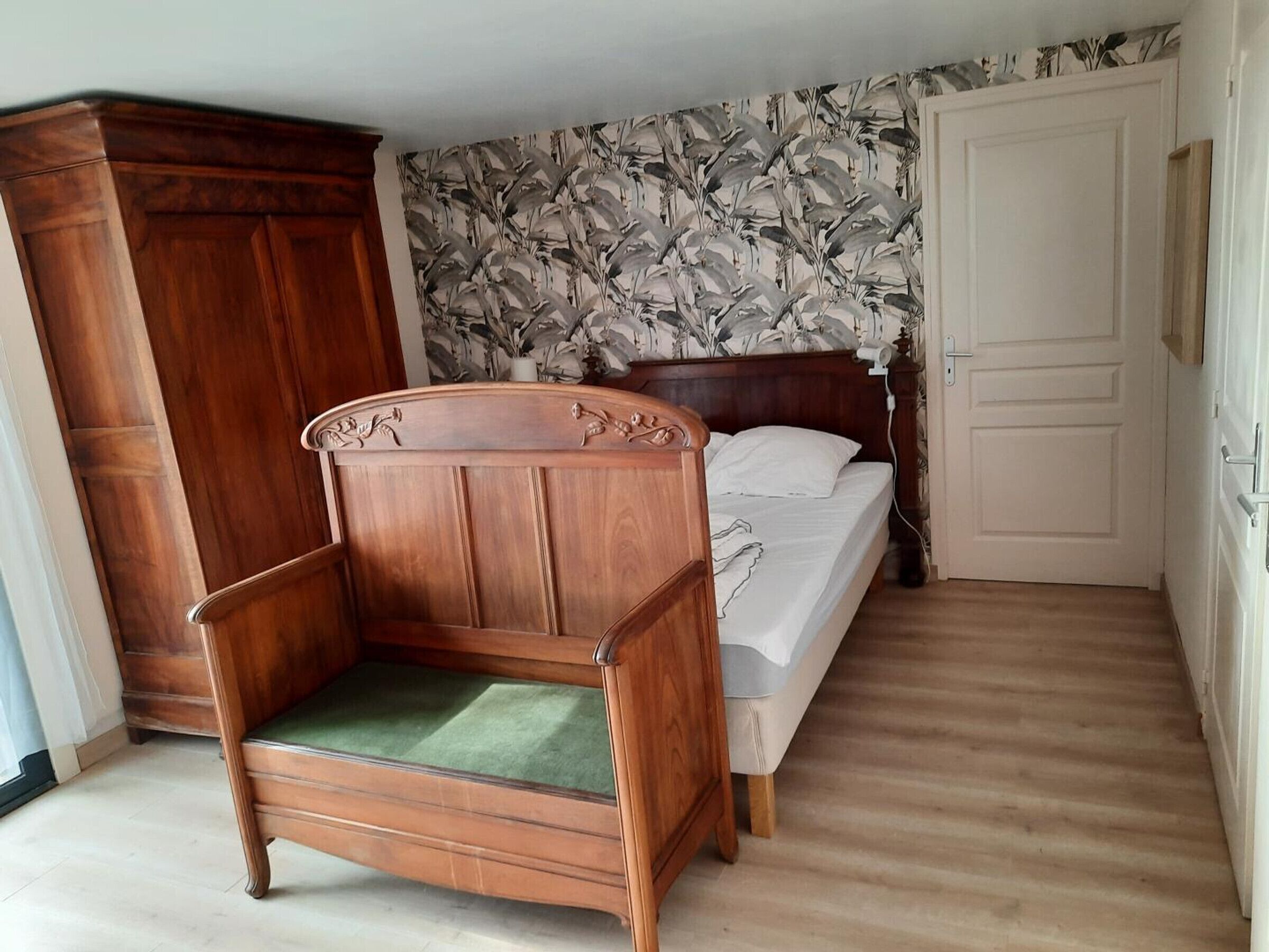Une chambre à coucher avec un lit en bois, un fauteuil recouvert d’un tissu vert, une armoire et un papier peint à motifs floraux.