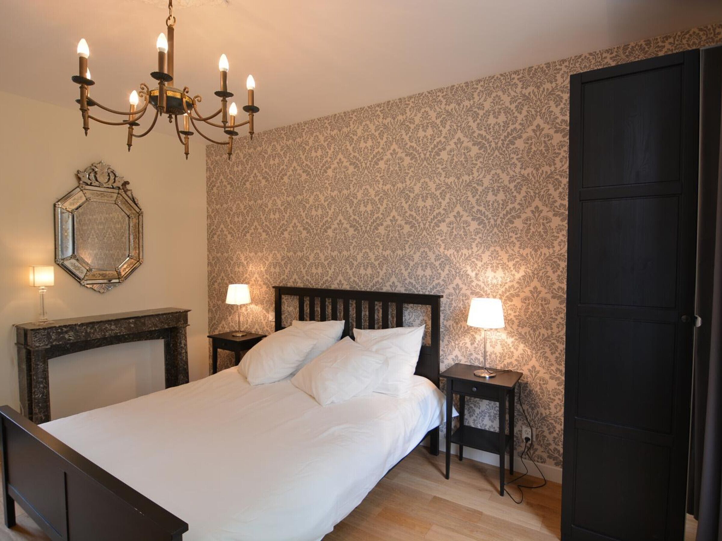 Un lit bien fait, avec du linge de lit blanc, une tête de lit en bois foncé et un lustre à plusieurs lampes. Il y a deux tables de chevet avec des lampes, et un grand miroir au mur.