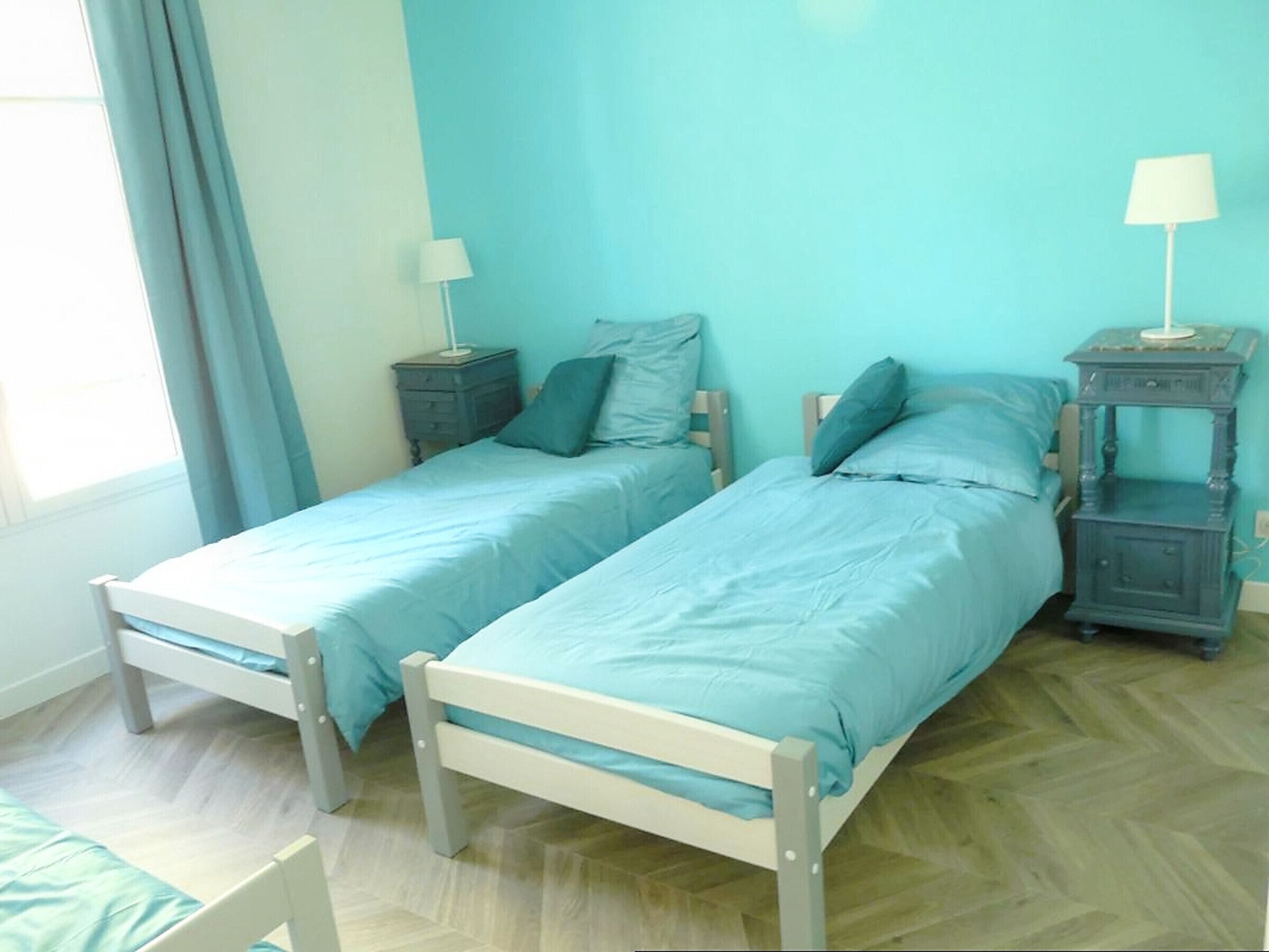 Une chambre dotée de deux lits simples, chacun avec une table de chevet et une lampe. La chambre a un parquet disposé en point de Hongrie et des murs de couleur bleu clair.