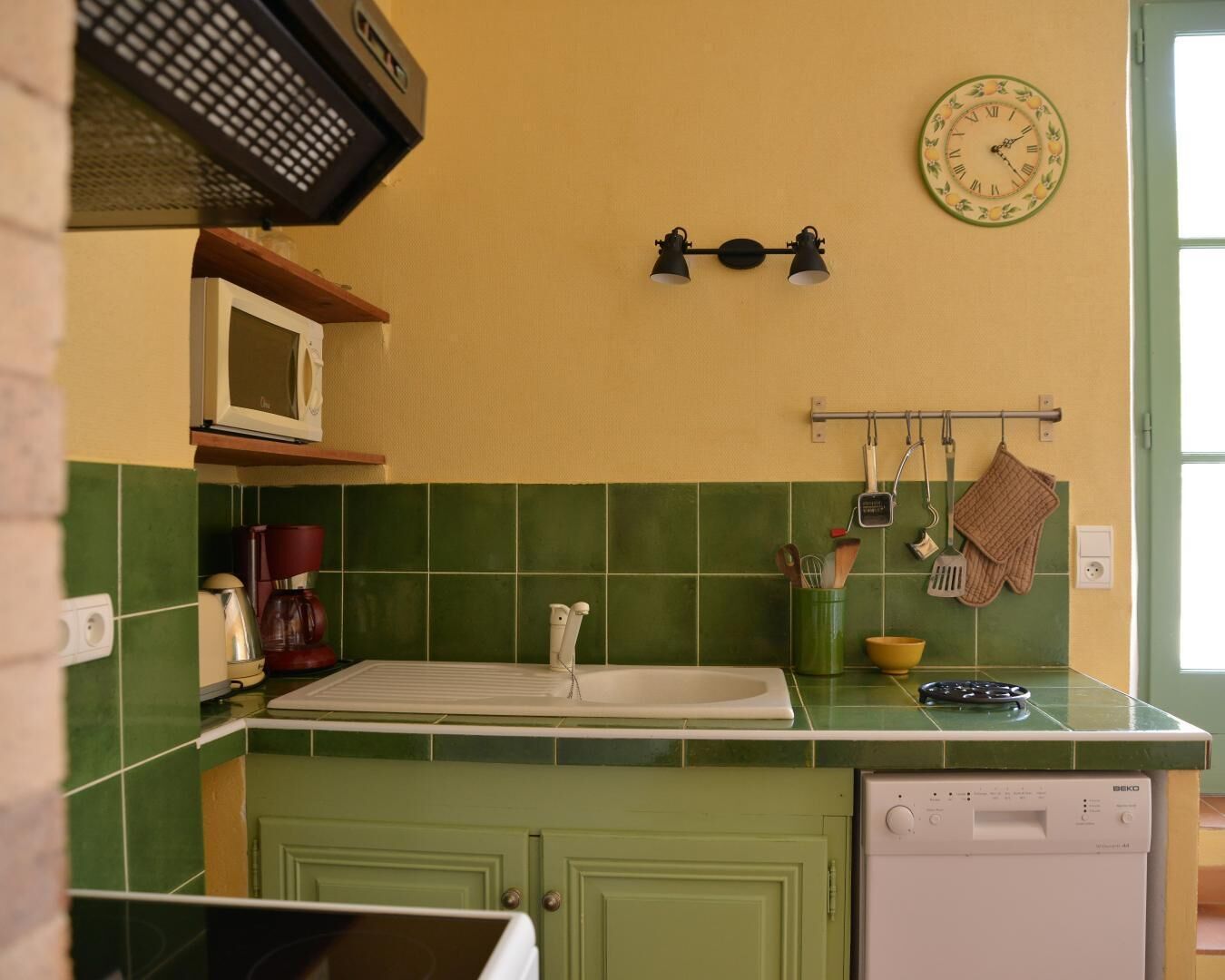 Een keuken met een groene betegelde achterwand, een witte wastafel, een magnetron en een klok aan de muur.