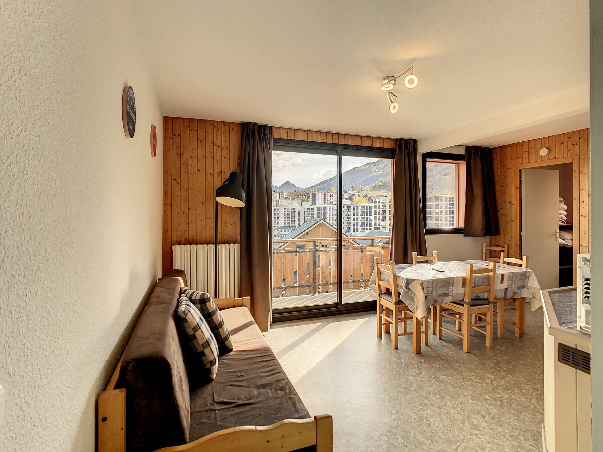 Une pièce chaleureuse avec un canapé en bois, une table à manger avec des chaises, une grande fenêtre donnant sur des bâtiments et des montagnes, et un luminaire au plafond.