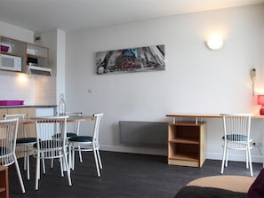 A compact living space with a kitchenette, a dining area with white chairs, a gray sofa, and a wall-mounted painting.