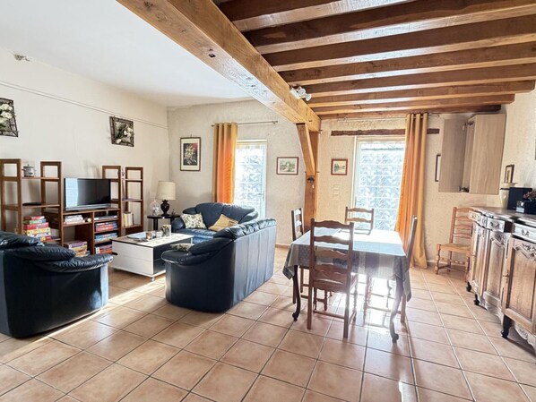 Un salon chaleureux avec un canapé, une table basse et un téléviseur, des poutres en bois au plafond et un coin repas avec une table et des chaises.