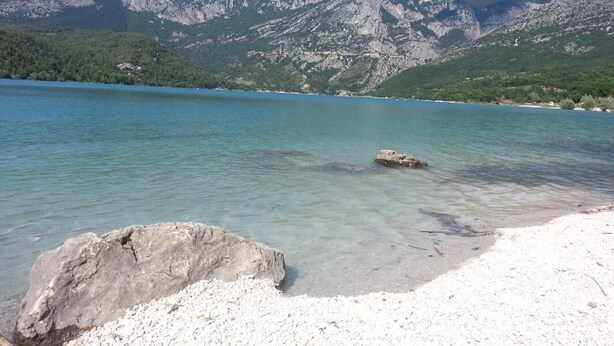 Un lac aux eaux limpides, bordé d’une rive rocheuse, avec des montagnes en arrière-plan.