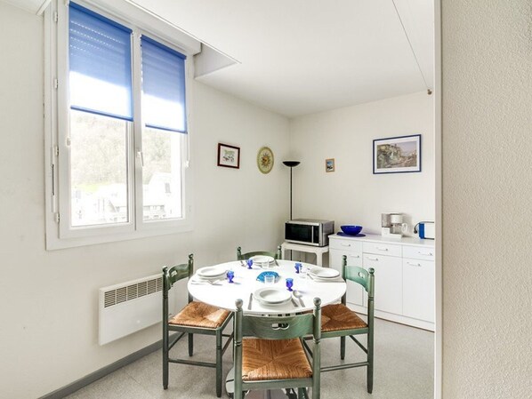 Un comedor con una mesa redonda dispuesta para cuatro personas, rodeada de sillas verdes con cojines marrones. Una ventana con persianas azules deja entrar luz natural, y hay un microondas visible en la encimera.
