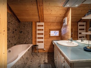 Een houten badkamer met een bad, twee wasbakken, een klein krukje en een raam met houten kader.