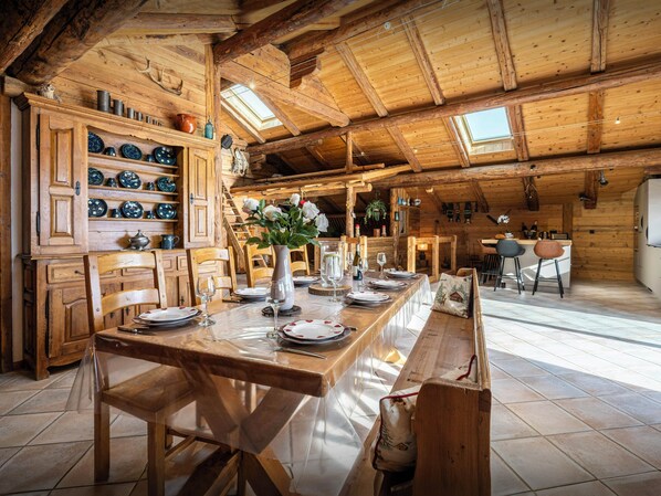 Een houten eethoek met een lange tafel, gedekt voor een maaltijd, houten kastjes met servies en een rustiek plafond met dakramen.