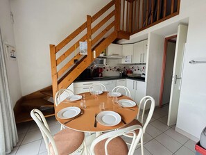 Un espace repas avec un escalier en bois, des chaises blanches et une cuisine en arrière-plan.