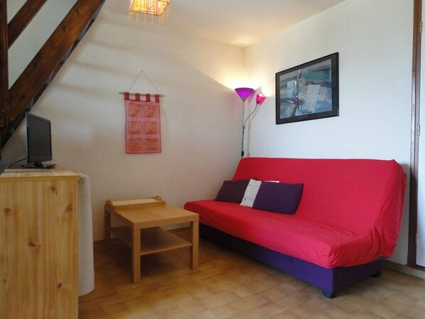 A cozy living room with a red sofa, a small wooden table, a TV on a wooden cabinet, a hanging pink banner, a floor lamp, and a painting on the wall.