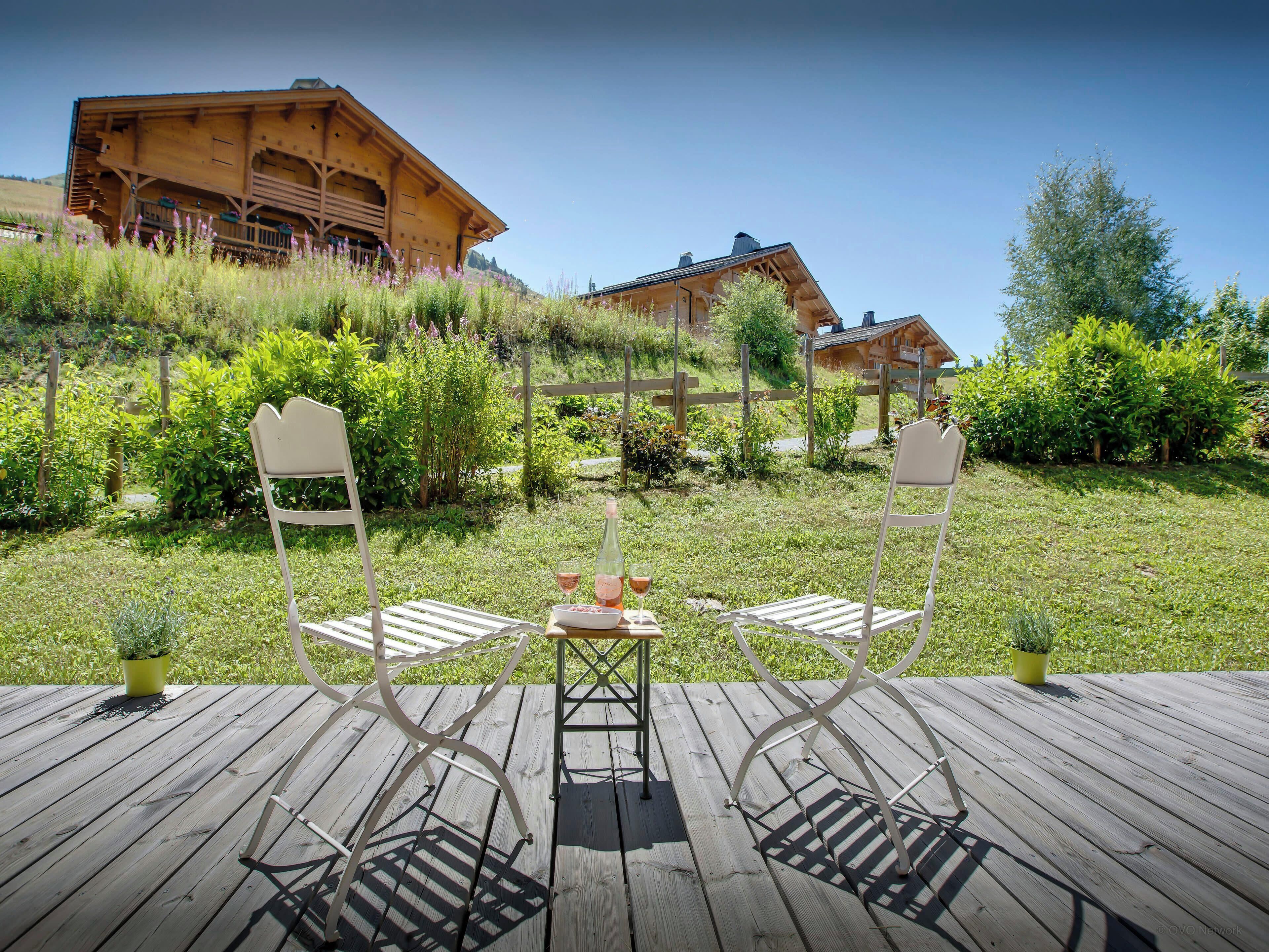 A wooden deck with two white chairs and a table, overlooking a lush garden and a wooden building.