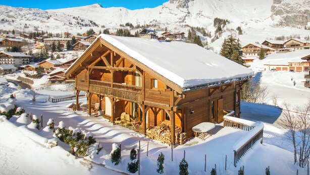 A wooden chalet with a balcony, surrounded by snow-covered mountains and other buildings.