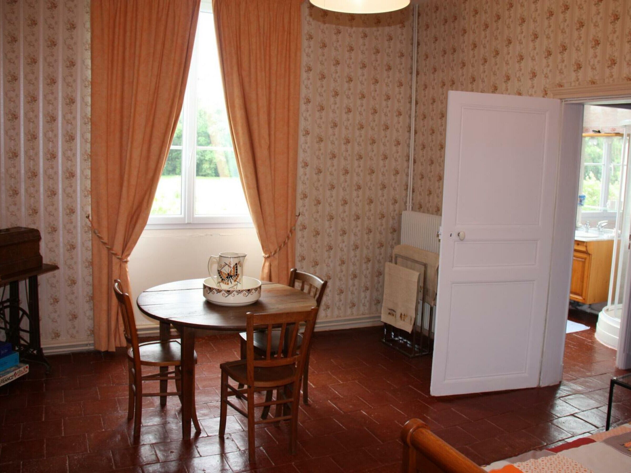 A dining area with a round table, four chairs, a pitcher, and a bowl. A window with curtains is visible, and a door leads to a kitchen.