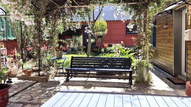 Un banco de madera bajo una pérgola con plantas colgantes, rodeado de un jardín con diversas macetas y un edificio rojo al fondo.