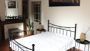 Un dormitorio ordenado con cama, una mesita, una silla, una lámpara colgante y un cuadro en la pared.