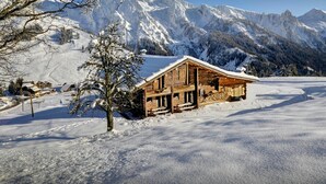 A wooden cabin with a snow-covered roof, surrounded by a snowy landscape and mountains.
