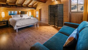 A wooden cabin room with a bed, a sofa, a nightstand, and a window with a view of mountains.