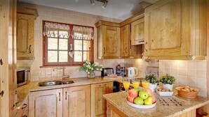A kitchen with wooden cabinets, a sink, a window with lace curtains, and a fruit bowl on the counter. - Chalet Chenavray - Manigod base for 10, spa & great skiing - OVO Network (Manigod)