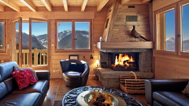 Interior acogedor de una cabaña de troncos con chimenea, sofás de cuero y vistas a montañas nevadas a través de amplias ventanas.