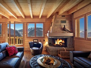 L’intérieur chaleureux d’un chalet en bois, avec une cheminée, des canapés en cuir et une vue sur des montagnes enneigées, visible à travers de grandes fenêtres.