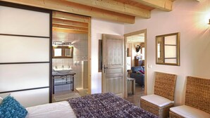 Un dormitorio con cama, una mesita de noche, un espejo, una silla de mimbre y un baño visible a través de una puerta abierta.
